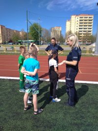 Dwie policjantki stoją przy boisku na terenie Ośrodka Sportu i rozmawiają z grupą chłopców w wieku 7-10 lat. Dzieci odwrócone tyłem do obiektywu