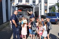 policjant z lewej strony, dzieci tyłem do zdjęcia, stoją przy radiowozie. Policjant pokazuje wyposażenie radiowozu.