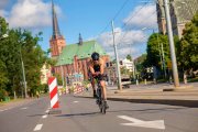 na zdjęciu widoczny mężczyzna jadący na rowerze, ubrany w sportowy kombinezon, okulary przeciwsłoneczne i kask. W tle widać budynek kościoła, drzewa.