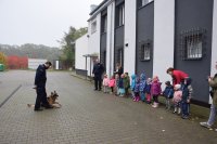 dzieci stoją przy budynku komendy po lewej stronie zdjęcia.  przed nimi po prawej stronie stoi policjant z psem służbowym.