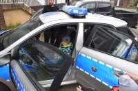 chłopiec siedzi w radiowozie na miejscu kierowcy, obok stoi policjant i pokazuje mu jak działają urządzenia zamontowane w pojeździe