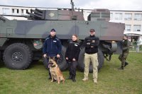 Na boisku szkolnym stoi funkcjonariusz policji z psem służbowym, uczennica klasy policyjnej i żołnierz, w tle stoi pojazd opancerzony &quot;Rosomak&quot;.