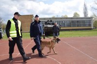 Przewodnik z psem służbowym idzie razem z uczniem z klasy policyjnej po boisku szkolnym w stronę radiowozu.