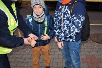 Policjantka stoi po lewej stronie, po prawej stronie dwóch chłopców. Policjantka zakłada opaskę odblaskową na rękę chłopca.
